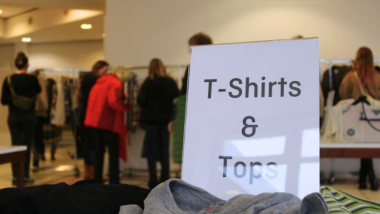 Im Vordergrund steht ein weißes Schild mit der Aufschrift "T-Shirts &amp; Tops", im Hintergrund stehen Personen mit dem Rücken zum Betrachter an Kleiderständern und schauen diese durch